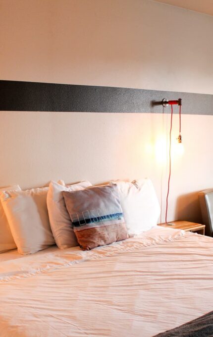 A neatly made bed with white linens and several pillows is positioned against a white wall with a horizontal dark stripe. The room features two chairs, a small table, a wall-mounted light, and framed artwork. The atmosphere is cozy and minimalist.