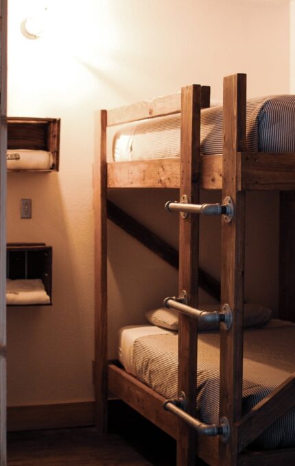 The image shows a small bedroom corner with wooden bunk beds. Each bunk has visible ladders, and the walls are made of wooden panels. To the left, there is a floor lamp and part of a gray armchair. The room has a simple, rustic style.