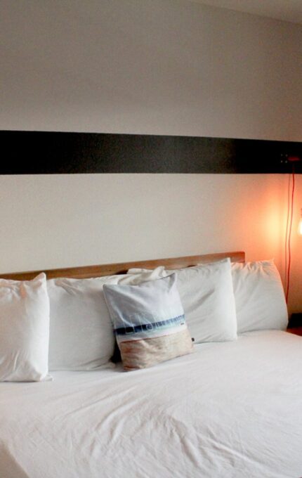 A bedroom with a large bed featuring white linens and multiple pillows, one with a striped design. Two Edison-style hanging light bulbs flank the headboard, casting a warm glow. The window on the right has a dark curtain partially drawn, letting in natural light.