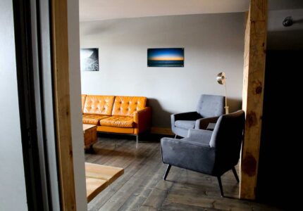 A living room with a gray wall, featuring an orange sofa and two gray armchairs arranged around a wooden coffee table. A tall, modern floor lamp is placed beside one armchair. Two landscape photographs hang on the wall. The floor is dark wooden.