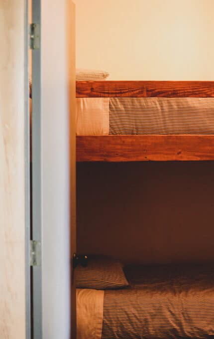 A vertical image shows a wooden bunk bed with two levels. Each bed has a mattress covered in striped bedding. The room appears to be minimally decorated, with the edge of a doorframe visible on the left side of the image.