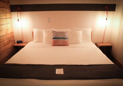 A neatly made bed with white linens, four pillows, and a gray blanket at the foot. A small note is placed on the bed. Bedside tables with lamps flank the bed. The wall behind features a horizontal dark stripe, and a digital clock is visible on the left side.