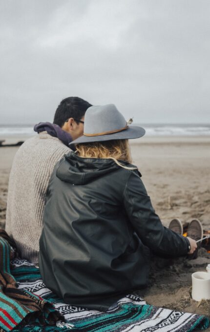 Two people sit on a striped blanket by a small campfire on a sandy beach. The person in the foreground wears a gray hat and a green jacket. There are two white mugs on the blanket. The ocean and overcast sky are visible in the background.