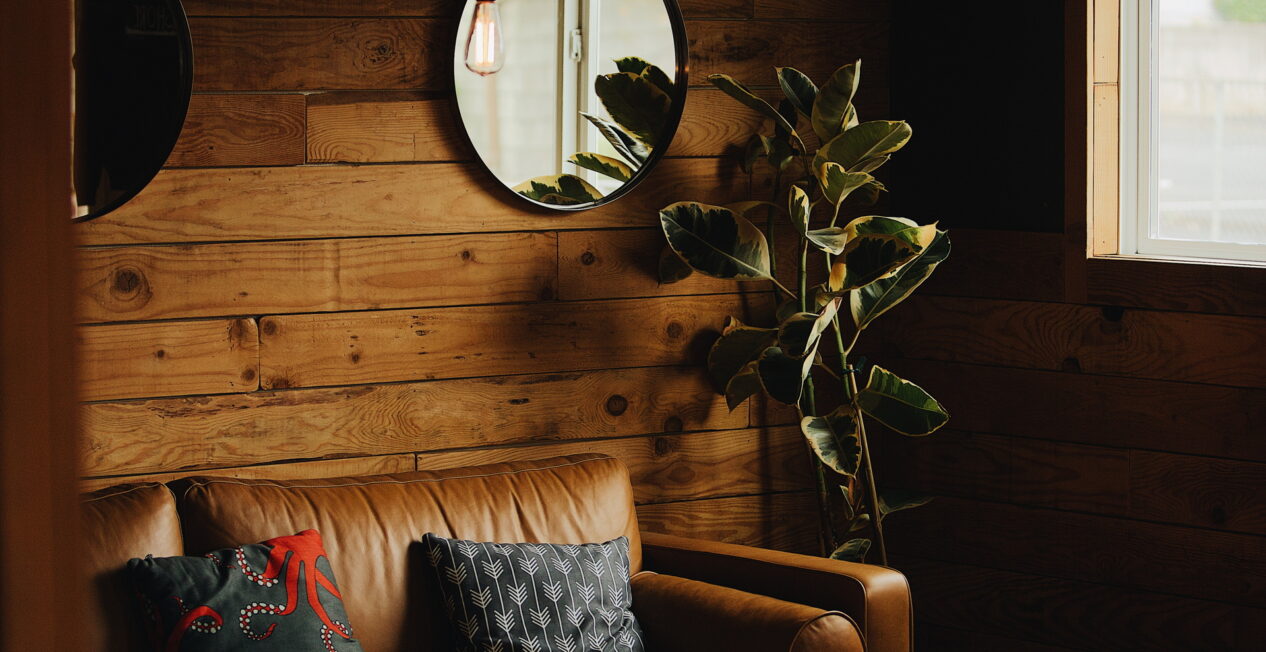 A cozy living room features a tan leather sofa with two patterned throw pillows against a wooden paneled wall. A round mirror and a potted plant adorn the space. A small round marble coffee table sits in front of the sofa, next to a window.