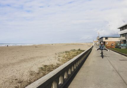 Person wearing a helmet rides a bicycle on a pathway adjacent to a sandy beach, lifting the front wheel in a wheelie. The beach is mostly empty, with a few people in the distance. Buildings are visible along the path, and the sky is partly cloudy.