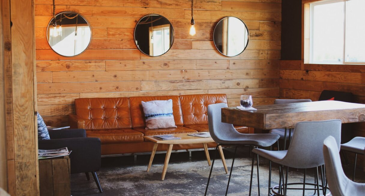 A cozy, modern living area with wooden walls featuring three round mirrors. It includes a brown leather couch, a small wooden coffee table, a dark armchair, and a high wooden table with four light gray stools. A window allows natural light into the room.