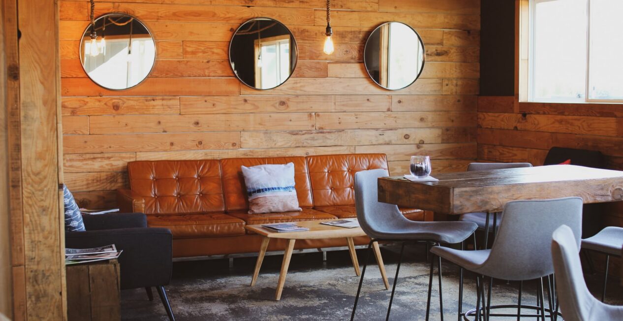 A cozy, modern living area with wooden walls featuring three round mirrors. It includes a brown leather couch, a small wooden coffee table, a dark armchair, and a high wooden table with four light gray stools. A window allows natural light into the room.