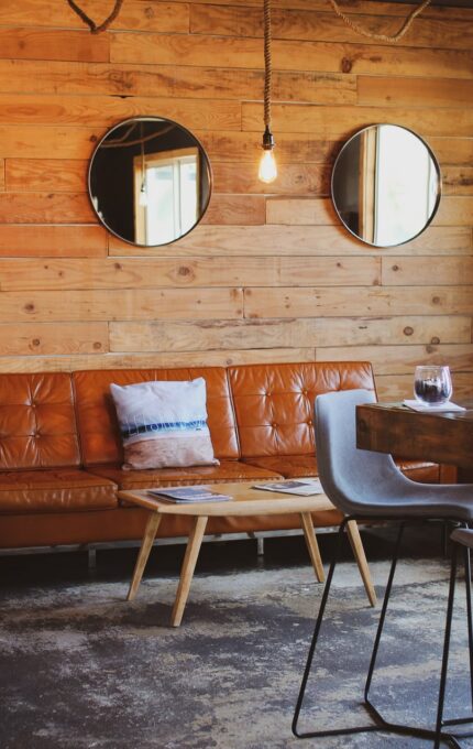 A cozy, modern living area with wooden walls featuring three round mirrors. It includes a brown leather couch, a small wooden coffee table, a dark armchair, and a high wooden table with four light gray stools. A window allows natural light into the room.