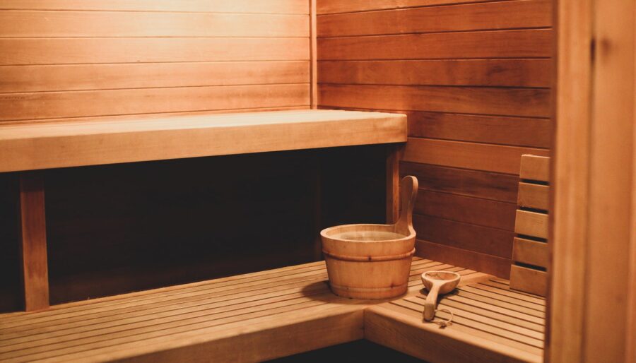 A wooden sauna interior featuring an L-shaped bench, wooden bucket, and ladle. The walls and benches are made of light-colored wood, and the space is warmly lit.