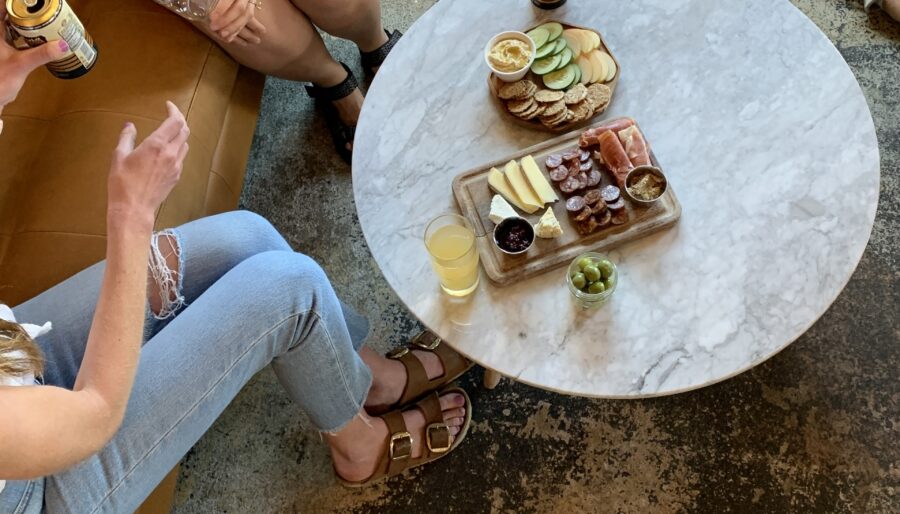 Two people sitting around a round marble table, each holding a drink. The table has a tray with assorted snacks like cheese, crackers, grapes, and sliced cucumbers. There is a third person’s foot visible in the upper right corner of the image.