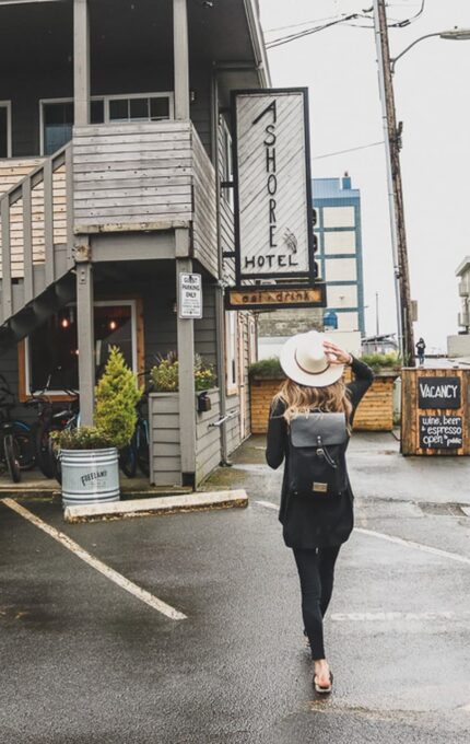 A person carrying a backpack and holding a hat walks toward a building labeled "Shore Hotel." The street is wet, indicating recent rain. Bicycles are parked under a staircase, and a vintage blue truck is in the distance. A sign shows "Vacancy" for the hotel.