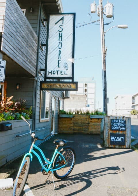 A blue bicycle is parked at the side of a road next to a hotel with a sign reading "Ashore Hotel." A chalkboard sign on the sidewalk advertises wine, beer, espresso, and room vacancy. A wooden building and utility pole are visible in the background.