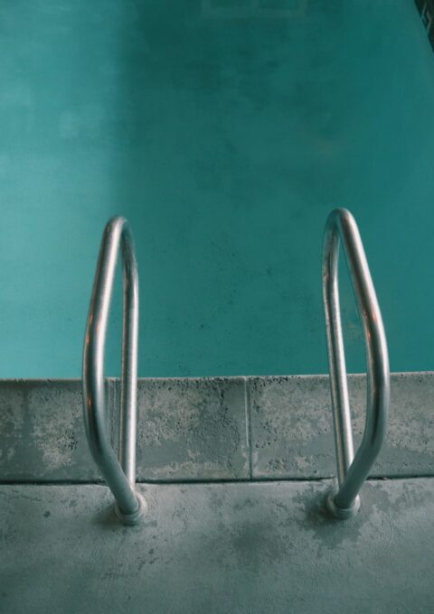 A corner of a swimming pool with a metal ladder is shown. The pool's water is turquoise, and the deck surrounding the pool is made of concrete with visible texture. The image is slightly darkened, giving it a shaded or early morning appearance.