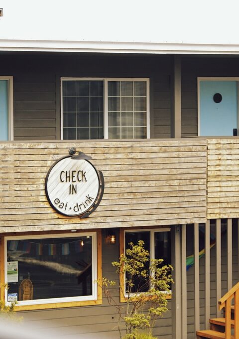 A two-story building with wooden paneling features a circular sign that reads "Check In, Eat, Drink" on the balcony railing. The second floor has light blue doors with small square windows. A staircase leads to the upper level, and there are some small trees nearby.