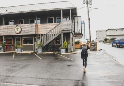 A person with a hat and backpack walks towards a two-story building with wood siding on a rainy day. The building has a sign that reads "Astoria Hotel" along with a "Vacancy" sign. Several bicycles are parked near the entrance.