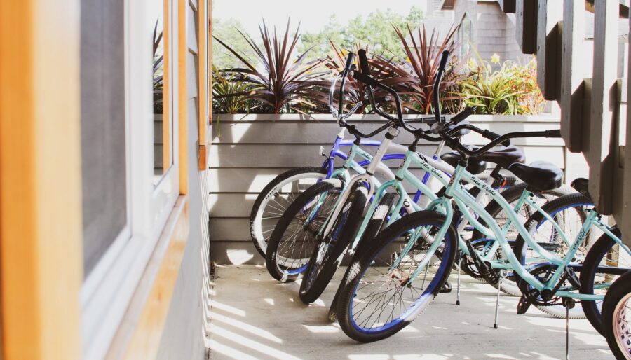 Several bicycles are parked in a row on a sunny outdoor patio next to a building. The bikes have colorful frames and are positioned against a planter box with green plants.