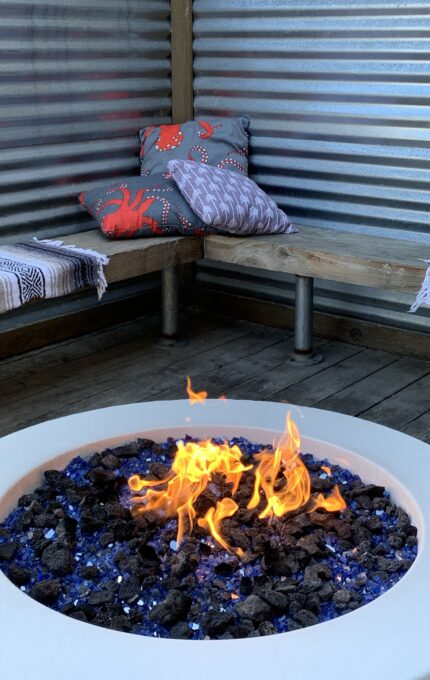 A round outdoor fire pit with flames burning surrounded by blue glass rocks. Behind it is a wooden bench covered with multicolored cushions and a corrugated metal wall.