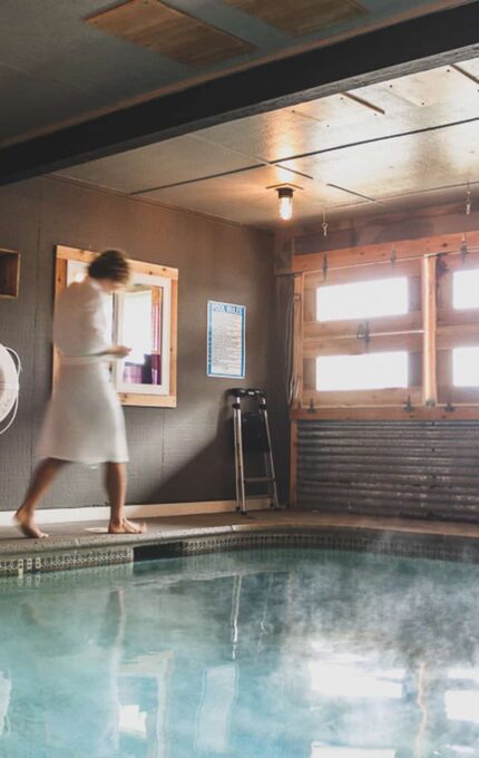 A person in a white bathrobe walks beside an indoor swimming pool. The room has wooden walls with a window, two red chairs, a lifebuoy, and shelves holding towels. Steam rises from the pool, and the space is lit by ceiling lights.