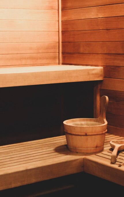 A wooden sauna room with benches on two sides, a bucket of water with a ladle placed in the corner, and wooden panels covering the walls and floor. The lighting is warm, giving the room a cozy and inviting atmosphere.