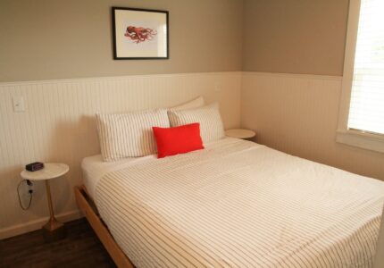 A small bedroom with a neatly made bed featuring white and gray striped bedding and a red accent pillow. There are two small round side tables beside the bed, one with a digital clock. A window with closed blinds and a framed picture are on the walls.