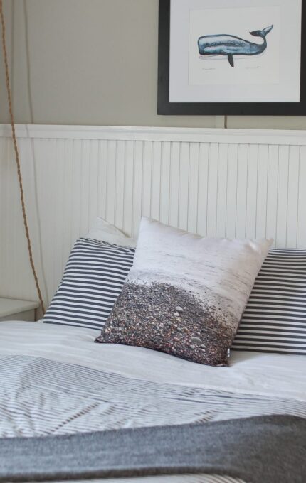 A neatly made bed with striped bedding, a pillow featuring a beach scene, and a gray blanket at the foot. A framed whale picture hangs on the light gray wall above the white beadboard wainscoting. A hanging light with an exposed bulb is on the left.