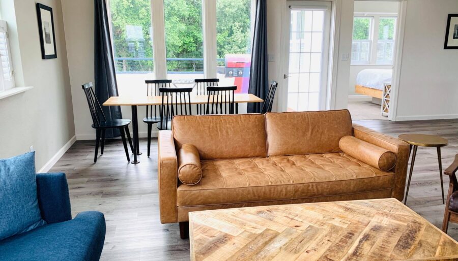 A living room features a brown leather sofa, a blue upholstered armchair, and a wooden coffee table with a geometric pattern. Behind the sofa, there is a dining area with a wooden table and black chairs. Large windows and a door lead to an outdoor area.