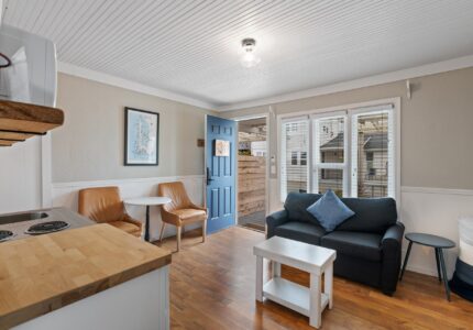 A cozy living space with wooden floors features a dark blue sofa with a cushion, two brown leather chairs, a small round table, and a white coffee table. The blue front door is open, leading to an outdoor area. A wall map and stove are also visible.