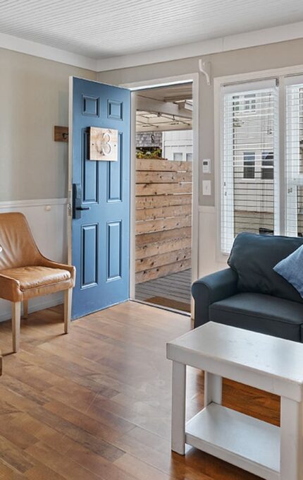 A cozy living area with a small kitchen on the left, featuring wooden countertops. There are two brown leather chairs and a small round white table. A dark blue couch with a blue pillow sits beside a white coffee table. The blue front door is open, revealing an outside view.