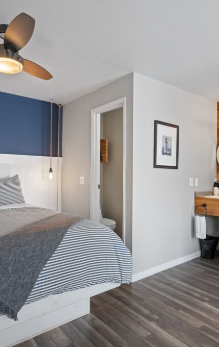 A cozy bedroom features a large bed with gray bedding, a ceiling fan, and a blue accent wall. The adjacent kitchenette includes a microwave, mini-fridge, and coffee maker. There is a small bathroom visible through a doorway. The room has wooden flooring and minimalist decor.