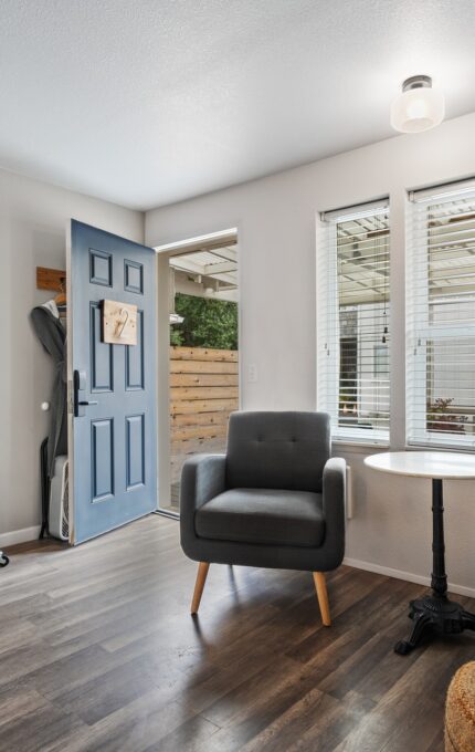 A small living area with two gray armchairs, a round white side table, and a wicker ottoman. A blue front door is ajar, revealing a wooden exterior. Kitchen appliances, including a microwave and mini-fridge, are visible on a wooden counter to the left.