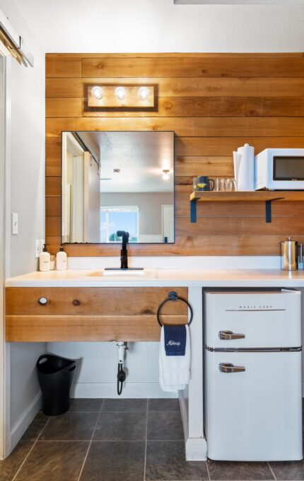 Modern bathroom with light wood paneling and white walls. To the left is a toilet with a shelf above it. The vanity has a wood countertop, black faucet, and large mirror. On the right shelving unit are a microwave and mini fridge, along with a coffee maker.