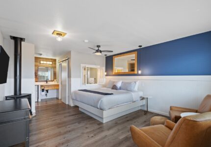 A modern hotel room features a bed with white and blue bedding, a dark blue accent wall, a large mirror, and a mounted TV. There is a seating area with two brown leather chairs and a side table. The bathroom with a well-lit vanity is visible in the background.