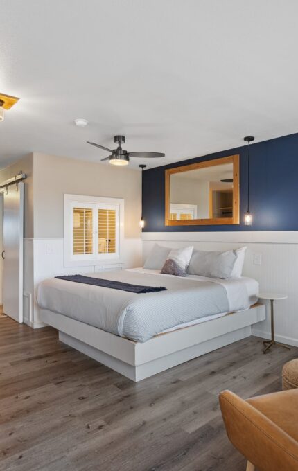 A modern bedroom features a raised bed with grey and white bedding, two leather armchairs, a wooden floor, a wall-mounted TV, a ceiling fan, and a blue accent wall. The room also includes an open bathroom with a wooden vanity and a sliding barn door.