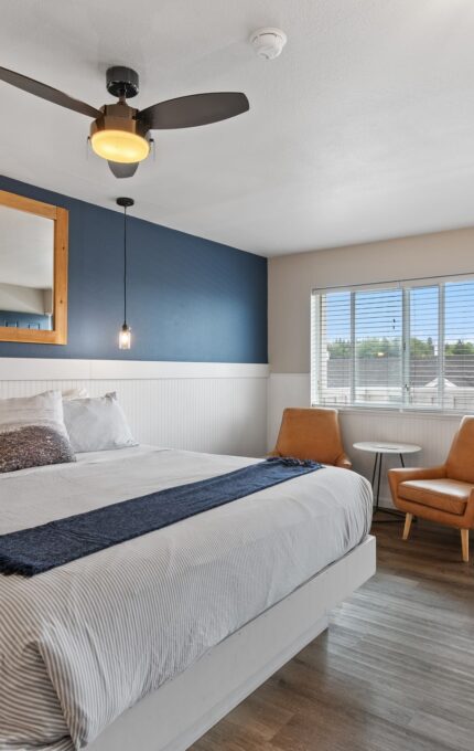 A modern bedroom with a large bed featuring white bedding and a dark blue throw. The room has a blue accent wall with a mirror, two pendant lights, two tan chairs with a small round table, a window, and a door leading to a balcony. The floor is wooden.