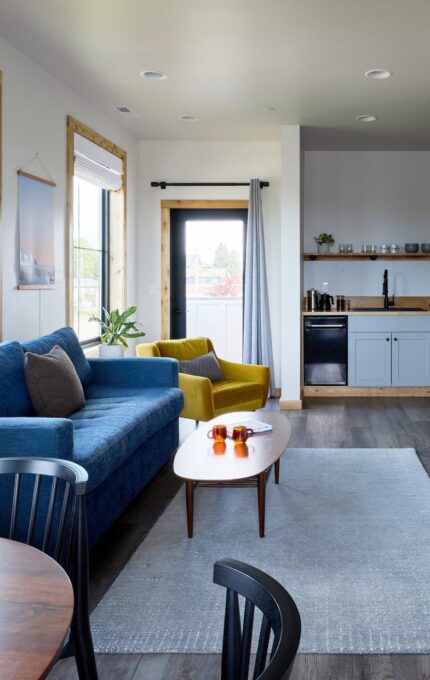 A modern living room with a blue sofa, yellow armchair, and coffee table. There is a round dining table with black chairs. The room features large windows, a wall-mounted TV, a fireplace, and an open kitchenette with a stove and shelving.