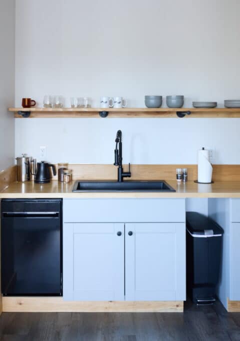 A modern kitchen features light gray cabinets, a black sink with a tall faucet, and a wooden countertop. Above, a wooden shelf holds mugs, bowls, and glasses. A black dishwasher, a toaster, and a kettle are placed on the countertops, along with a paper towel holder.