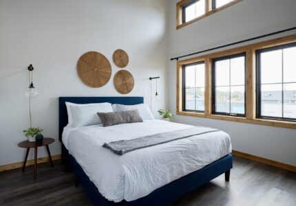 A modern bedroom with a large bed featuring white bedding and a dark blue frame. The room has wooden floors, white walls, and a tall ceiling. Three wicker wall decorations are hung above the bed. There are windows on two walls allowing natural light in.