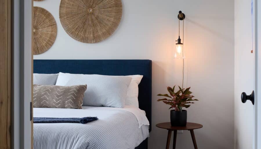 A neatly made bed with white and blue bedding stands against a white wall adorned with woven circular decorations. To the right, a wooden side table holds a potted plant and a hanging light fixture glows softly. The floor is wood, and a wooden door frame is visible.