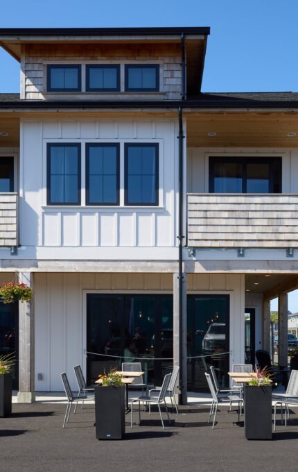 Two-story white building with a wooden roof and black-framed windows. Outdoor seating with tables and chairs is set up on the paved area in front. A sign near the entrance indicates an area of interest. The sky is clear and blue.