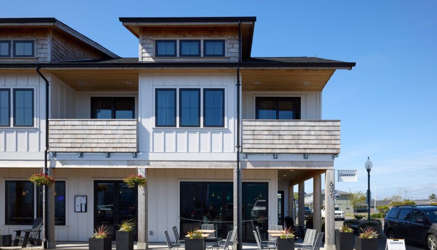 Two-story white building with a wooden roof and black-framed windows. Outdoor seating with tables and chairs is set up on the paved area in front. A sign near the entrance indicates an area of interest. The sky is clear and blue.