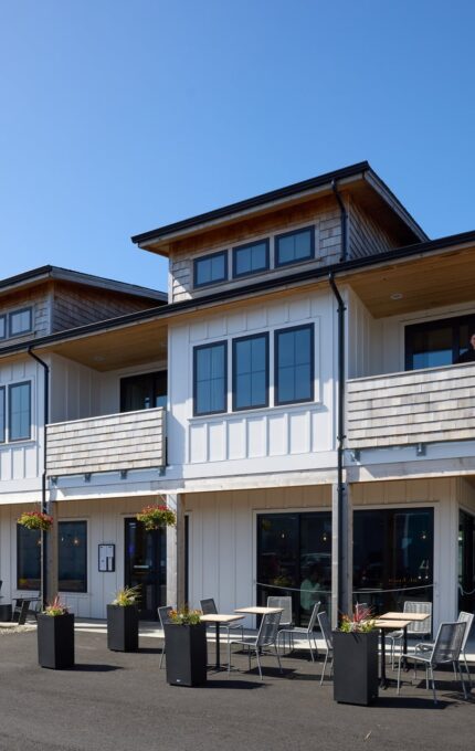 A modern, two-story building houses a restaurant on the ground floor. The exterior features white paneling with wooden accents. Outdoor seating is arranged near the entrance, and a man stands on the balcony of the upper floor. A clear sky is in the background.