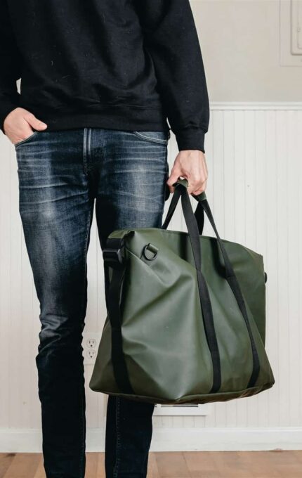 A person standing indoors, wearing a black sweatshirt and blue jeans, is holding a green duffel bag. The person has one hand in their pocket and the other holding the bag. The setting appears to be a room with white paneling and hardwood floors.