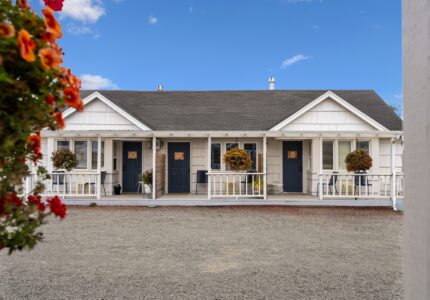 Image shows a single-story building with three front doors and small porches. Each porch has a potted plant or hanging flower basket. The building has white siding and a dark roof. In the foreground is a gravel driveway and a partially visible hanging basket with flowers.