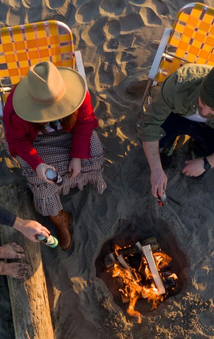 A top-down view of three people sitting in orange and yellow folding chairs around a small campfire on a sandy beach. One person holds a drink can, while another is adjusting a log on the fire. A log bench and some books or magazines are also visible.