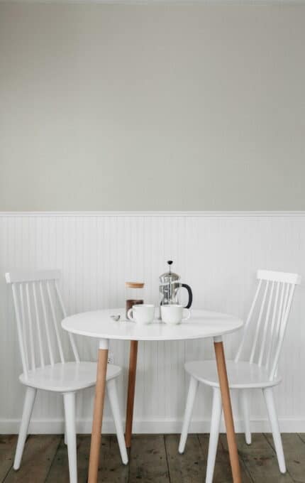 A small white round table with wooden legs is set with two white chairs, a French press, two white coffee cups, and a small saucer. The scene is on a wooden floor against a wall with white wainscoting and a light gray upper section.