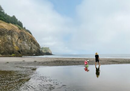 A person and a child walk along a sandy beach with shallow water reflecting their images. The beach is bordered by a rocky cliff with green foliage. The sky is partly cloudy, and the ocean is visible in the background.