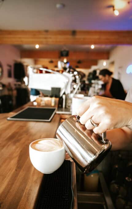 A barista's hand holding a metal pitcher is pouring steamed milk into a coffee cup on a wooden counter in a café. In the background, another person is visible working near coffee equipment. The café has a warm, inviting ambiance with exposed wooden beams.