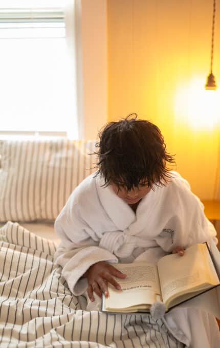 A person in a white bathrobe sits on a bed with striped bedding, reading a book. Warm lighting from a nearby lamp illuminates the scene.