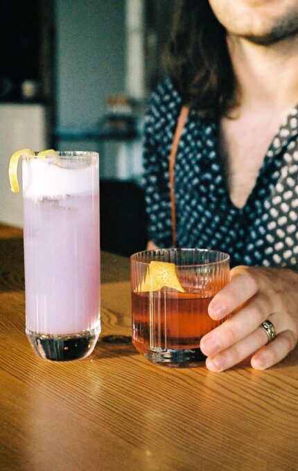 A person with long hair and a patterned shirt holds a glass of amber-colored cocktail with a lemon peel garnish, next to a tall glass of a light purple drink with ice and a lemon twist, on a wooden table.