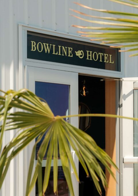 Entrance to Bowline Hotel, featuring a sign with the hotel's name and a bowline knot symbol above the door. White wall with a large glass door partially open. Palm leaves in the foreground frame the view.